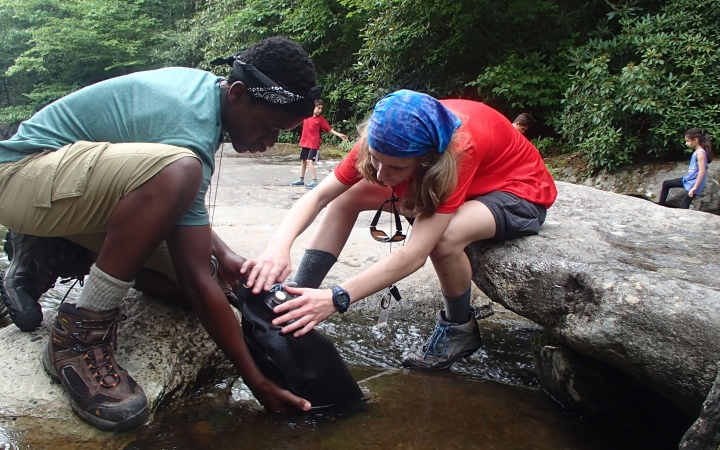 Two students kneel on rocks as they fill a water bladder in shallow water.